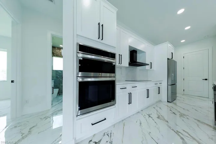 Kitchen featuring wall chimney exhaust hood, stainless steel appliances, recessed lighting, light marble finish flooring, and white cabinets
