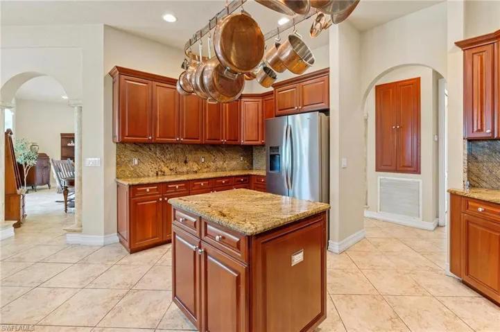 Kitchen with arched walkways, stainless steel fridge, decorative backsplash, light tile patterned flooring, and light stone counters