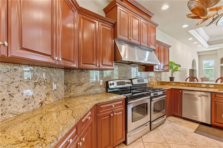 Kitchen featuring stainless steel appliances, under cabinet range hood, light stone countertops, backsplash, and light tile patterned flooring