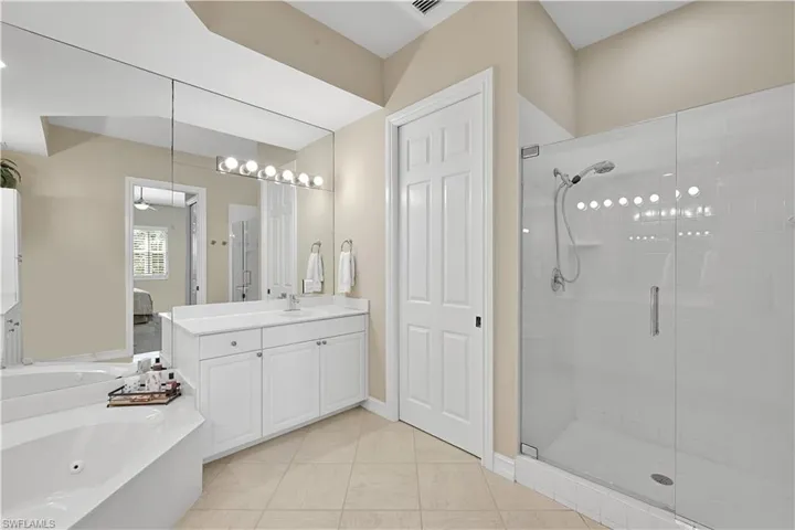 Bathroom featuring a white vanity with integrated basin, expansive wall-to-wall mirror, tiled soaking tub, walk-in shower with glass enclosure, and ceramic floor tile