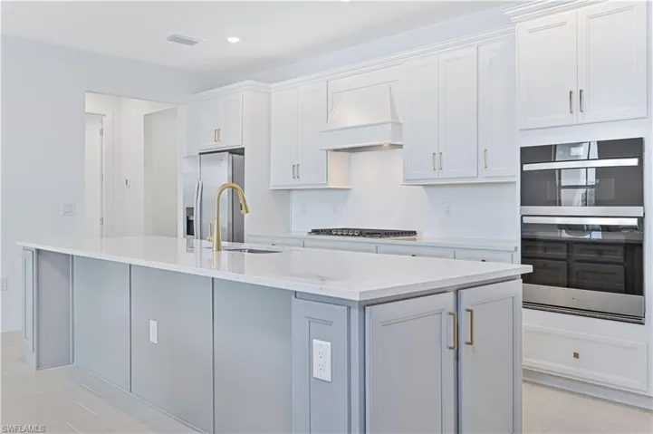 Kitchen featuring white cabinetry, appliances with stainless steel finishes, custom range hood, light stone countertops, and a center island with sink