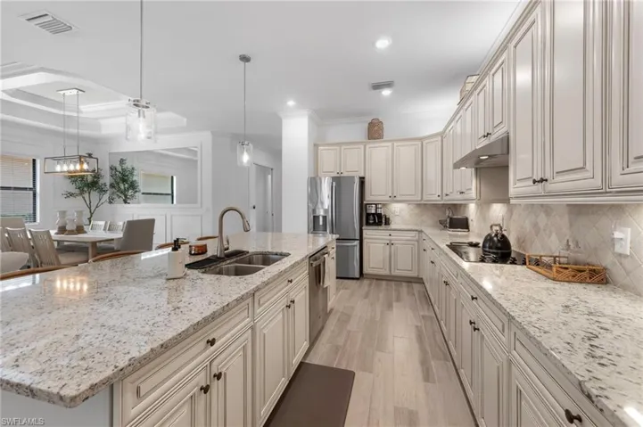 Kitchen with ornamental molding, light stone counters, hanging light fixtures, light wood-style floors, and an island with sink