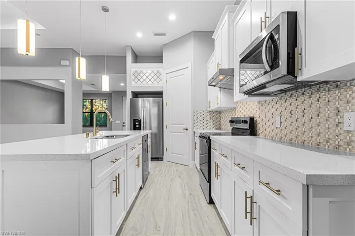 Kitchen with stainless steel appliances, white cabinets, an island with sink, light stone counters, and decorative light fixtures