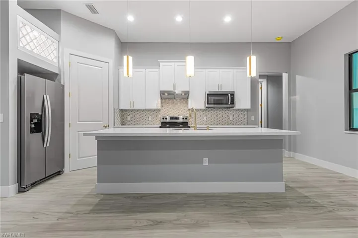 Kitchen featuring stainless steel appliances, white cabinetry, an island with sink, pendant lighting, and light stone countertops