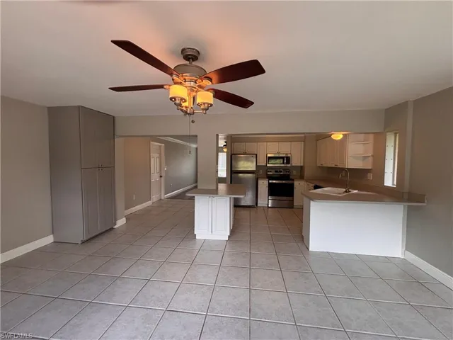 This open-concept living space features tiled flooring throughout, a ceiling fan with integrated lighting, and a kitchen area with white cabinetry, stainless steel appliances, and a kitchen island
