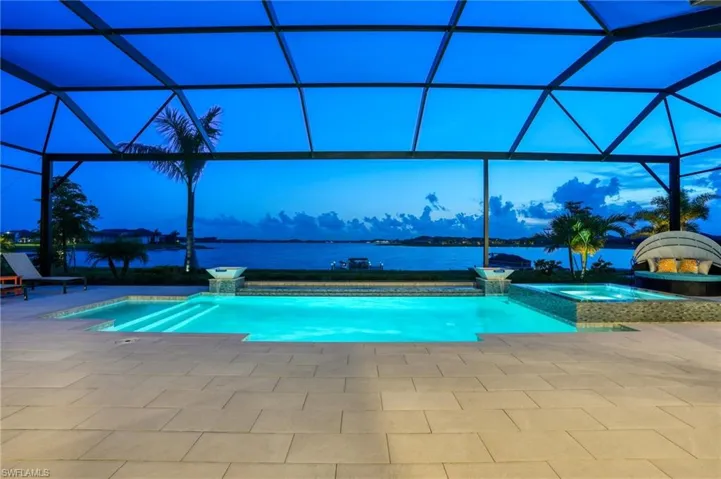 Pool at dusk featuring a water view, glass enclosure, a pool with connected hot tub, and a patio