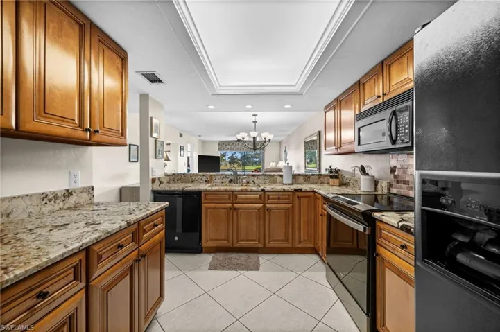 Kitchen featuring a chandelier, brown cabinetry, black appliances, light stone countertops, and a tray ceiling