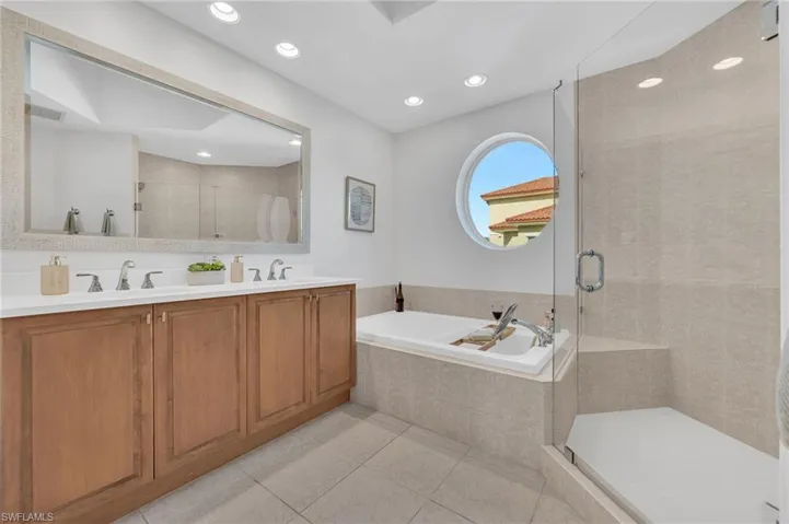 Bathroom featuring a double vanity with light wood cabinetry, a large frameless mirror, and a built-in tub with surrounding tile