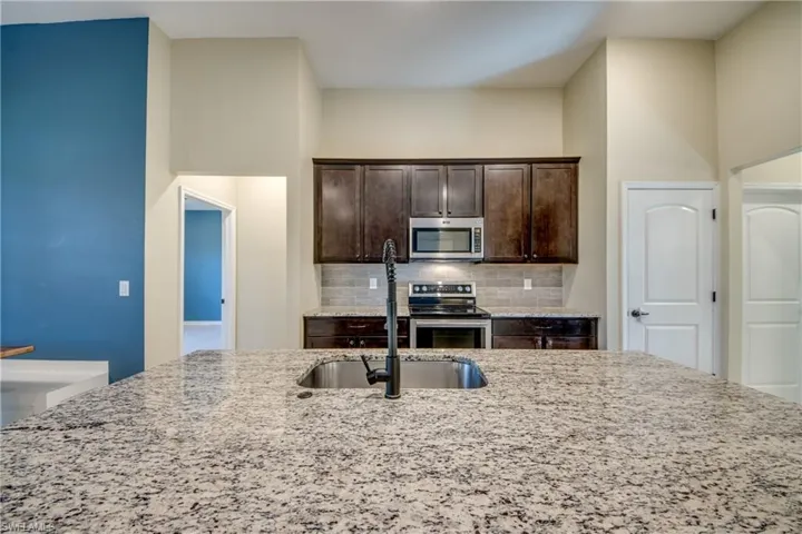 Kitchen featuring dark wood finish cabinets, light stone counters, stainless steel appliances, tasteful backsplash, and a high ceiling