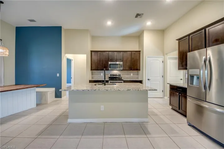 Kitchen featuring stainless steel appliances, dark wood finish cabinetry, light stone counters, tasteful backsplash, and a center island with sink