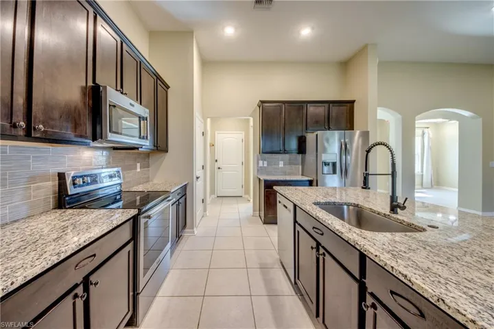Kitchen with stainless steel appliances, dark wood finish cabinets, arched walkways, light stone countertops, and backsplash