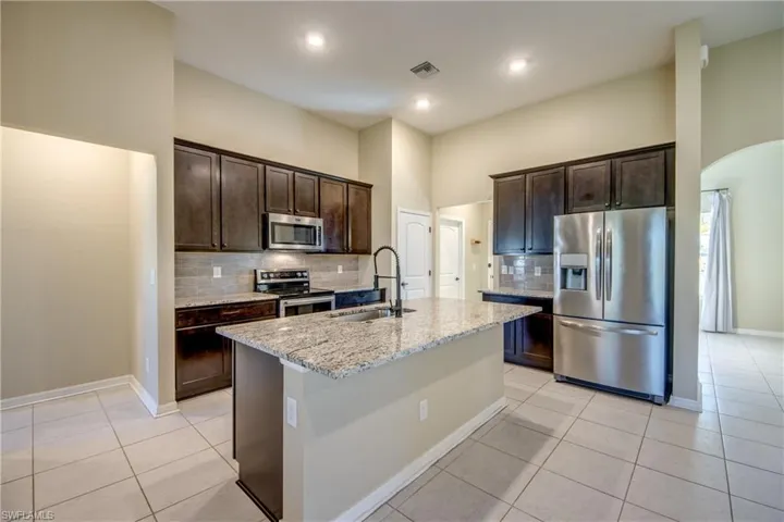 Kitchen featuring backsplash, stainless steel appliances, dark wood finish cabinets, light stone counters, and arched walkways