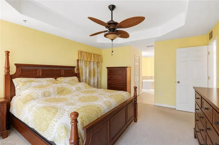 Virtually Staged Carpeted bedroom featuring a tray ceiling with a ceiling fan, light yellow painted walls, and a doorway opening to a tiled area with a built-in tub