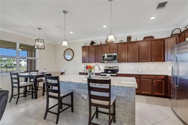 Kitchen with pendant lighting, stainless steel appliances, light stone counters, ornamental molding, and a kitchen island with sink