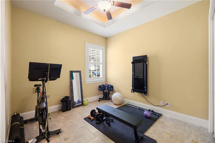 Exercise room featuring a raised ceiling, a ceiling fan, light stone finish flooring, and crown molding