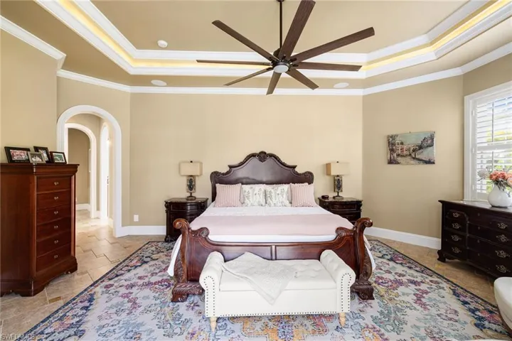 Bedroom featuring arched walkways, a raised ceiling, ceiling fan, crown molding, and stone tile flooring