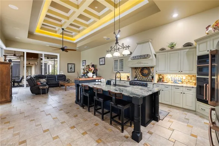 Dual tone kitchen with light stone counters, decorative light fixtures, a kitchen breakfast bar, high coffered ceiling, and an island with sink
