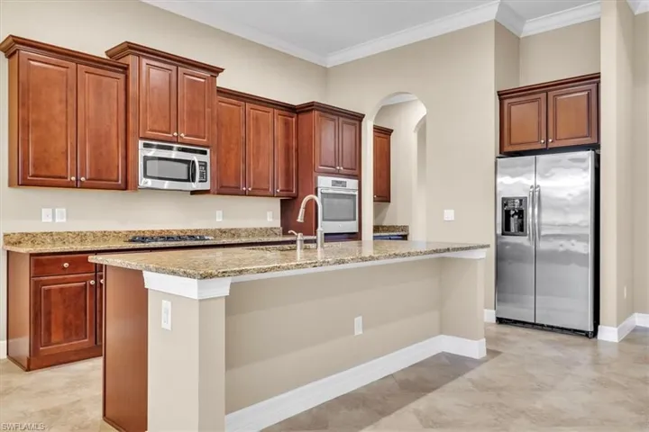 Kitchen featuring stainless steel appliances, light stone counters, arched walkways, an island with sink, and crown molding