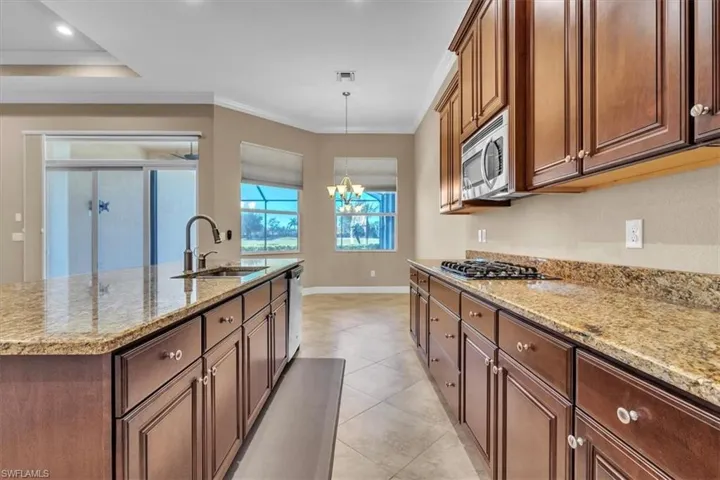 Kitchen with light stone counters, hanging lights, an island with sink, ornamental molding, and stainless steel appliances