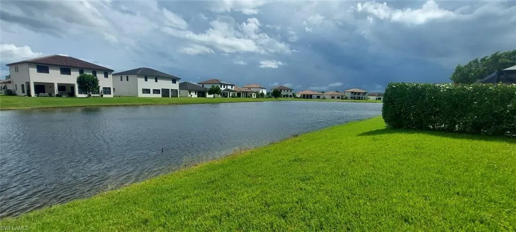 Expansive green lawn bordering a tranquil body of water, featuring a well-maintained hedge and residential structures with various rooflines in the background