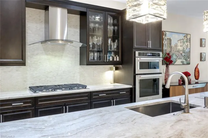 Kitchen with ventilation hood, stainless steel appliances, glass insert cabinets, light stone counters, and dark wood finish cabinetry