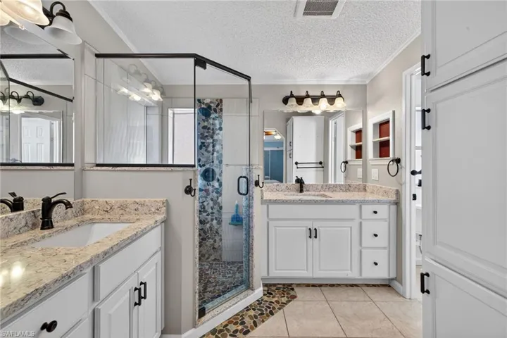 Primary Bathroom featuring a shower stall, a textured ceiling, two vanities, light tile patterned floors, and ornamental molding