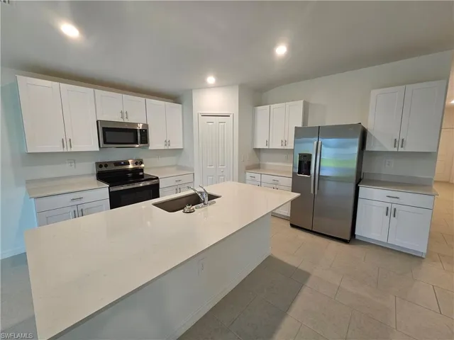 Kitchen featuring an island with an undermount sink, stainless steel appliances, white cabinetry, light-toned countertops, and recessed lighting
