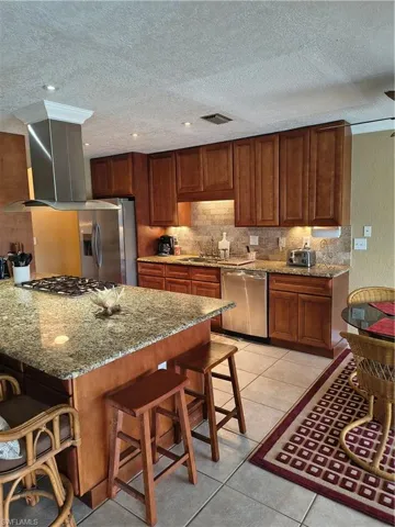 Kitchen featuring a granite-top island with cooktop, stainless steel range hood, ample wood cabinetry, mosaic tile backsplash, and stainless steel appliances