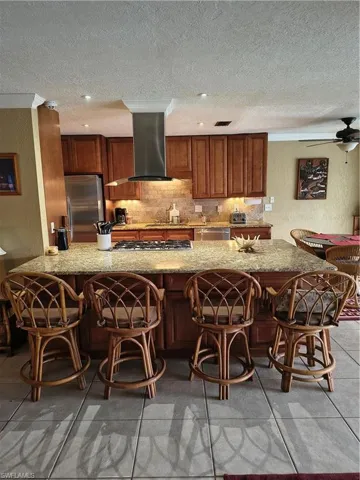 Kitchen featuring a large island with a built-in gas cooktop, stainless steel range hood, ample cabinetry, tile backsplash, and light-toned countertops