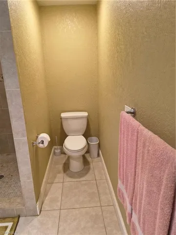 Private commode area featuring textured walls, tile flooring, a white toilet, and a chrome towel bar