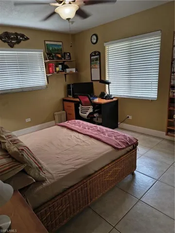 Bedroom featuring a decorative ceiling fan with integrated lighting, two windows with horizontal blinds, built-in wall shelving, and neutral tone tile flooring