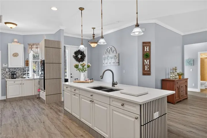 Kitchen featuring ornamental molding, hanging light fixtures, white cabinetry, light stone counters, and a kitchen island with sink