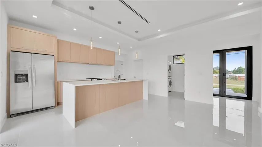 Kitchen with light brown cabinets, stainless steel fridge, decorative light fixtures, estacked washer and dryer, and an island with sink