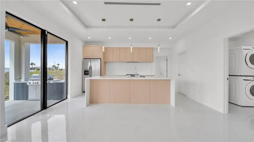 Kitchen featuring light brown cabinetry, pendant lighting, a center island with sink, stainless steel refrigerator with ice dispenser, and stacked washing machine and dryer