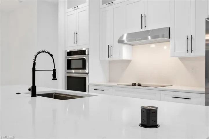 Kitchen featuring light stone countertops, white cabinetry, under cabinet range hood, double oven, and glass insert cabinets