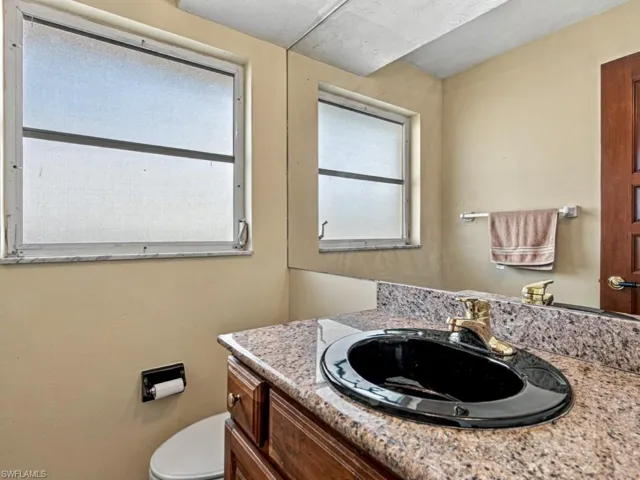 Vanity featuring stone-finish countertop, undermount sink with brass-finish faucet, wall-spanning mirror, and textured privacy window