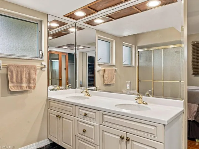 Double vanity featuring a solid surface countertop, dual integrated sinks, gold-tone fixtures, custom-framed cabinetry, and an expansive wall-mounted mirror with integrated wood-trimmed recessed lighting