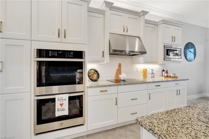 Kitchen with stainless steel appliances, under cabinet range hood, light stone countertops, white cabinetry, and crown molding