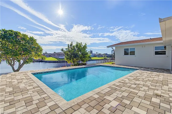 In-ground swimming pool surrounded by a light-toned paver patio, situated on a canal-front lot with mature landscaping