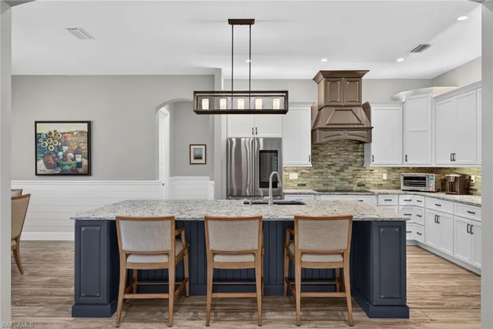 Expansive kitchen featuring an oversized island with a light-toned countertop, white cabinetry with dark hardware, a wood-finish range hood, a mosaic tile backsplash, and stainless steel appliances