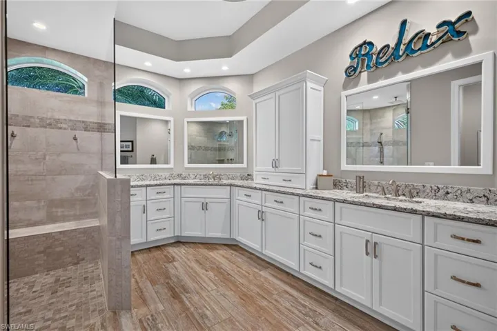 Primary Bathroom featuring wood-finish flooring, an expansive double vanity with white cabinetry and stone countertops, a large walk-in shower with tiled walls and floor, and arched transom windows