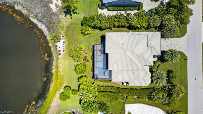 Aerial property perspective featuring a metal roof, screened pool enclosure, paved driveway, lush landscaping, and a lakefront setting