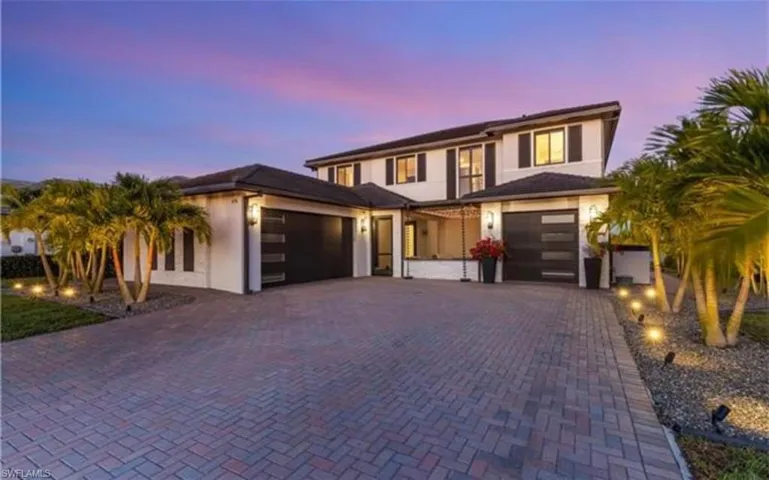 Contemporary exterior featuring a paver driveway, dual garage doors with frosted glass panels, mature palm trees, and exterior accent lighting