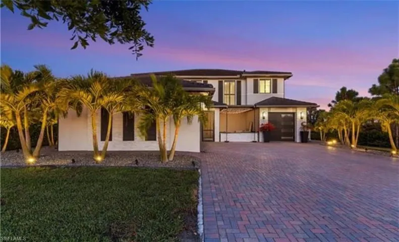 Two-story contemporary residence featuring a paver driveway, an attached garage, a stucco exterior, and accent lighting illuminating established palm trees