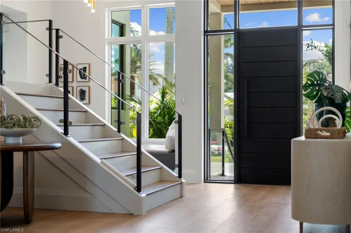 Wide plank wood-finish flooring, staircase with glass and black metal railing, and a black solid entry door with a large handle, complemented by expansive transom windows