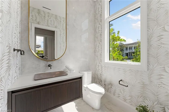 White window frame, white toilet, wall-mounted dark wood-finish vanity, gold-framed mirror, and patterned wallpaper