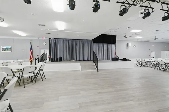 Open-concept hall featuring light-toned wood-finish flooring, a raised stage with curtain backdrop, and professional-grade overhead lighting fixtures