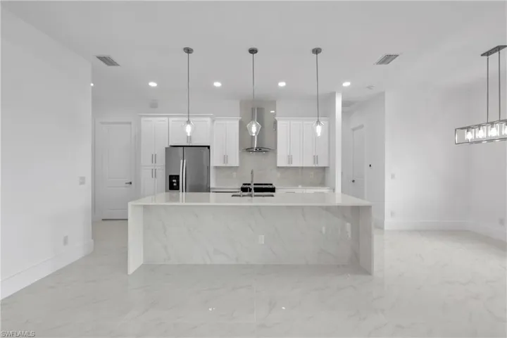 Kitchen with backsplash, wall chimney exhaust hood, stainless steel refrigerator with ice dispenser, an island with sink, and light tile patterned floors