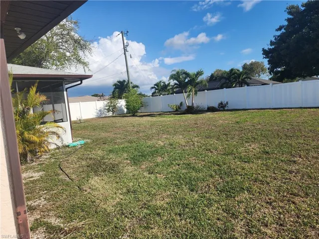 Expansive grassy yard area featuring a white privacy fence, mature palm trees, and a screened-in porch with a brown gutter system