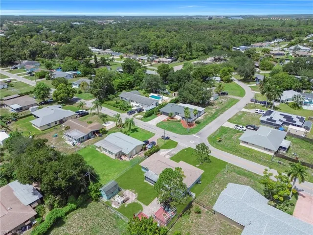 Aerial view showcasing the property's perimeter, featuring a single-story residence with a brown shingle roof, a concrete driveway, an expansive green lawn, and a paved patio area